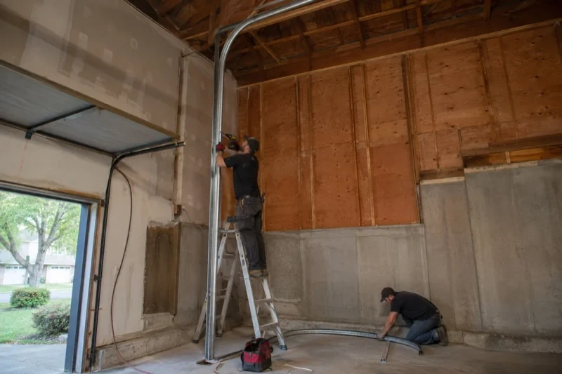 One person on a stepladder working beside a tall curved garage-door track while another kneels beside loose track sections in an unfinished garage.